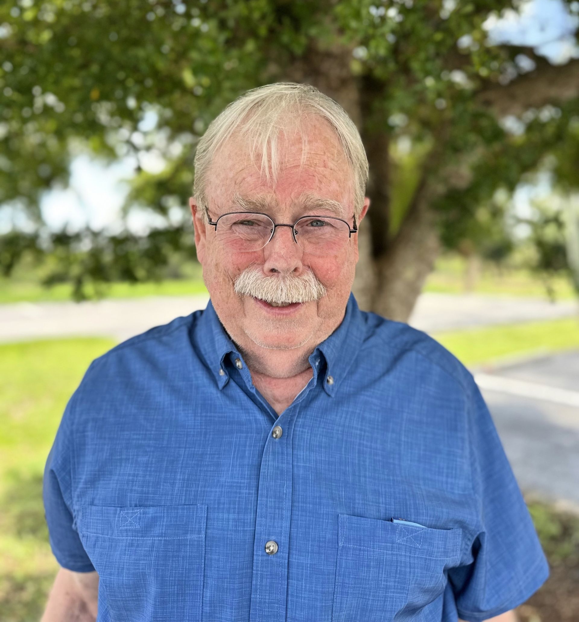 A man wearing glasses and a blue shirt is standing in front of a tree.