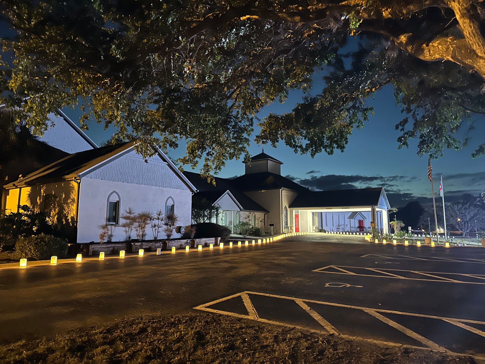A nighttime view of a building with lights and a tree canopy. The building is white and has a red door.