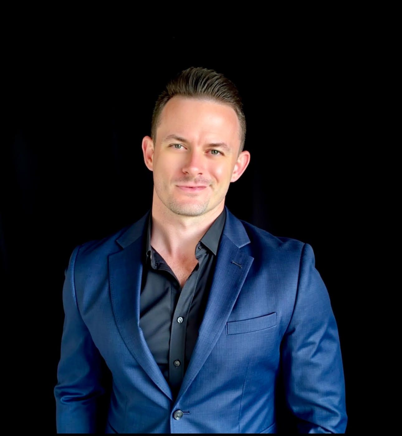 A man in a blue suit and black shirt is standing in front of a black background.