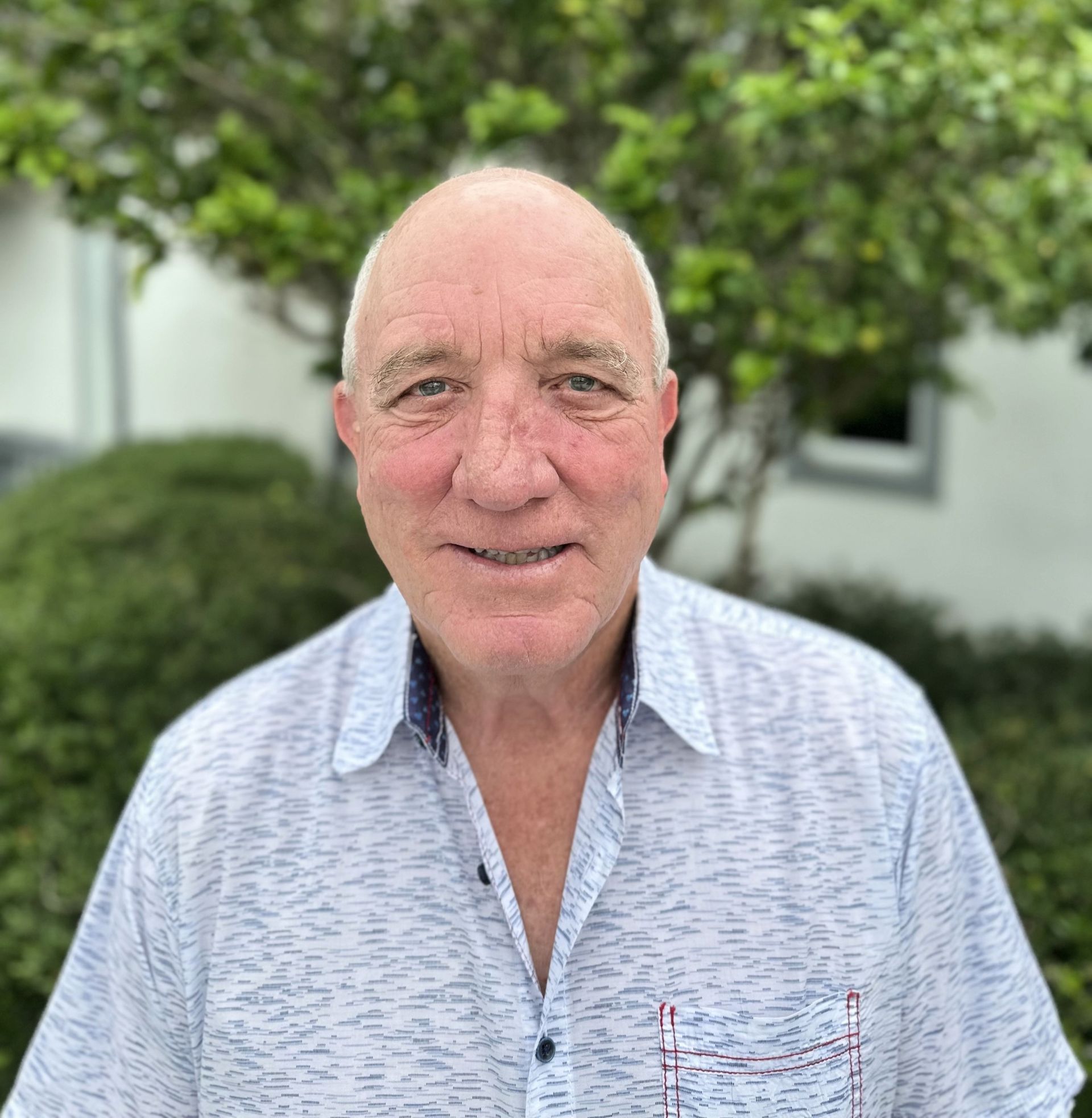 A man in a white shirt is smiling for the camera while standing in front of a tree.