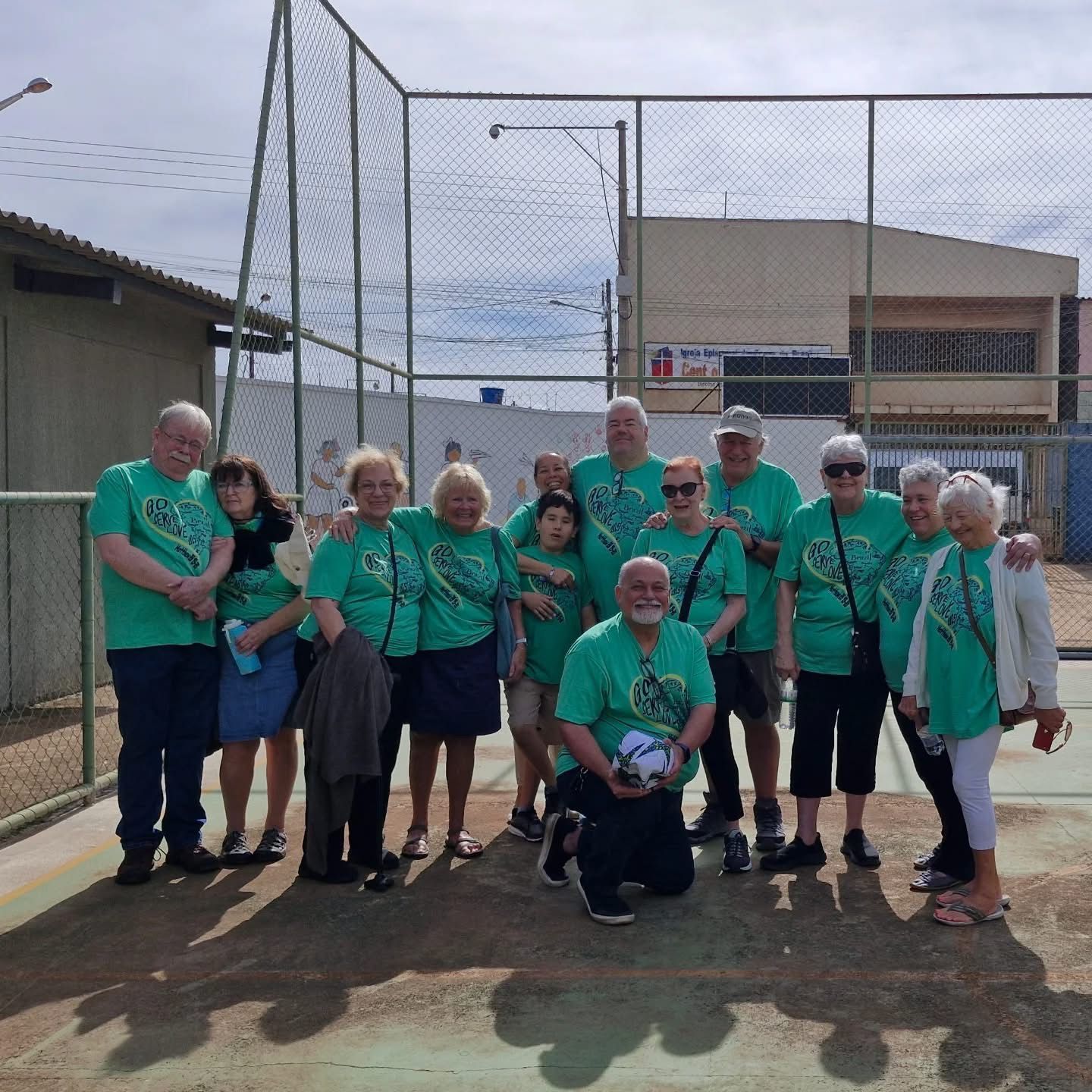 Group of seniors in green shirts pose with a soccer ball near a fence and building on a sunny day.