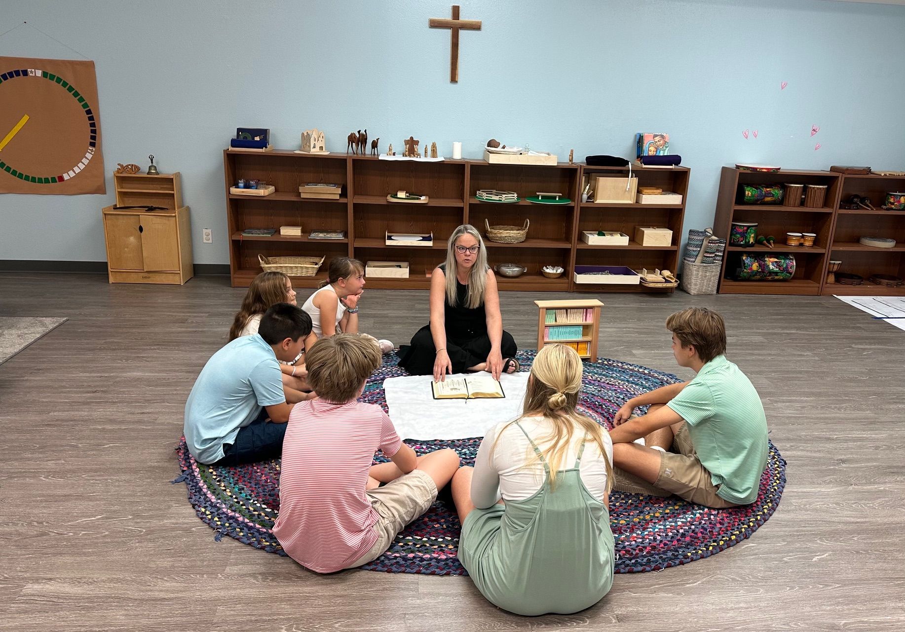 A teacher reads to children seated in a circle on a rug, shelves with materials in background, cross above.