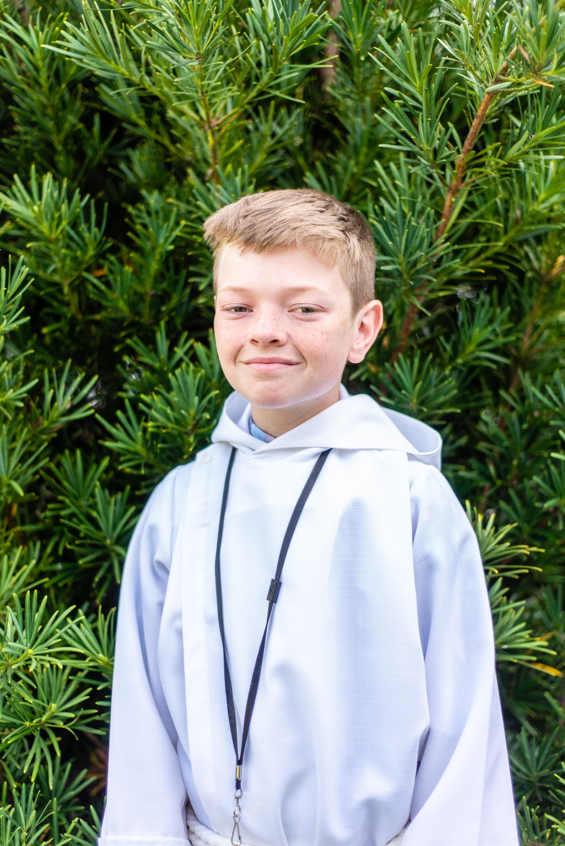 A young boy in a white robe is standing in front of a bush.
