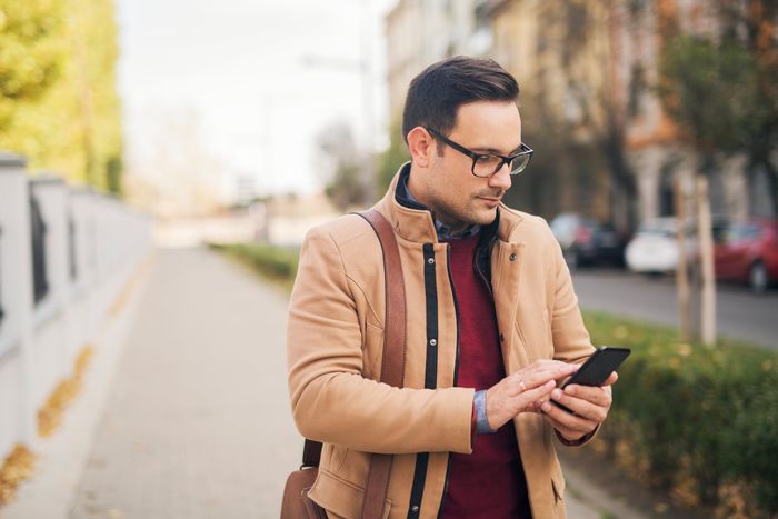 Young businessman with eyeglasses standing on the street and typing a message on his phone while waiting for limousine service in Westport, CT.