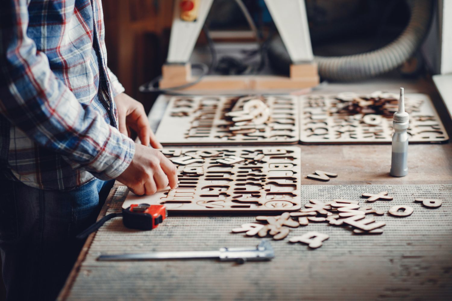 Una persona con una camisa a cuadros clasifica pequeñas letras de madera cortadas con láser en un banco de trabajo equipado 