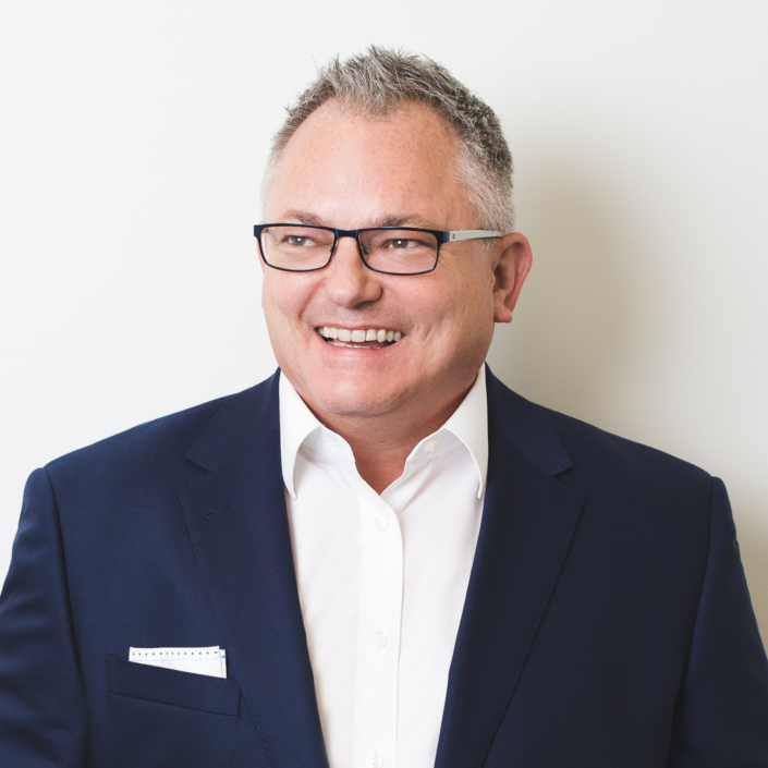 Man wearing glasses, white shirt, and navy blazer, smiling, in front of a white background.