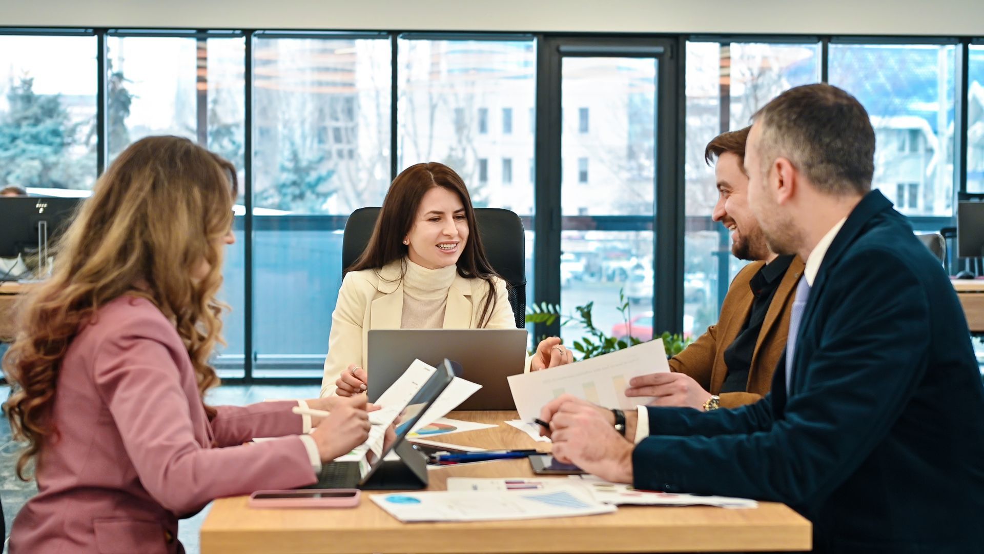 Four business professionals sit around a table in a modern office, collaborating on documents and a laptop during a meeting.