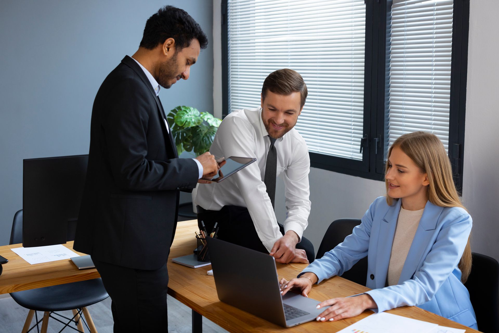 Three professionals collaborate in an office, reviewing content on a laptop and a tablet while standing around a desk.