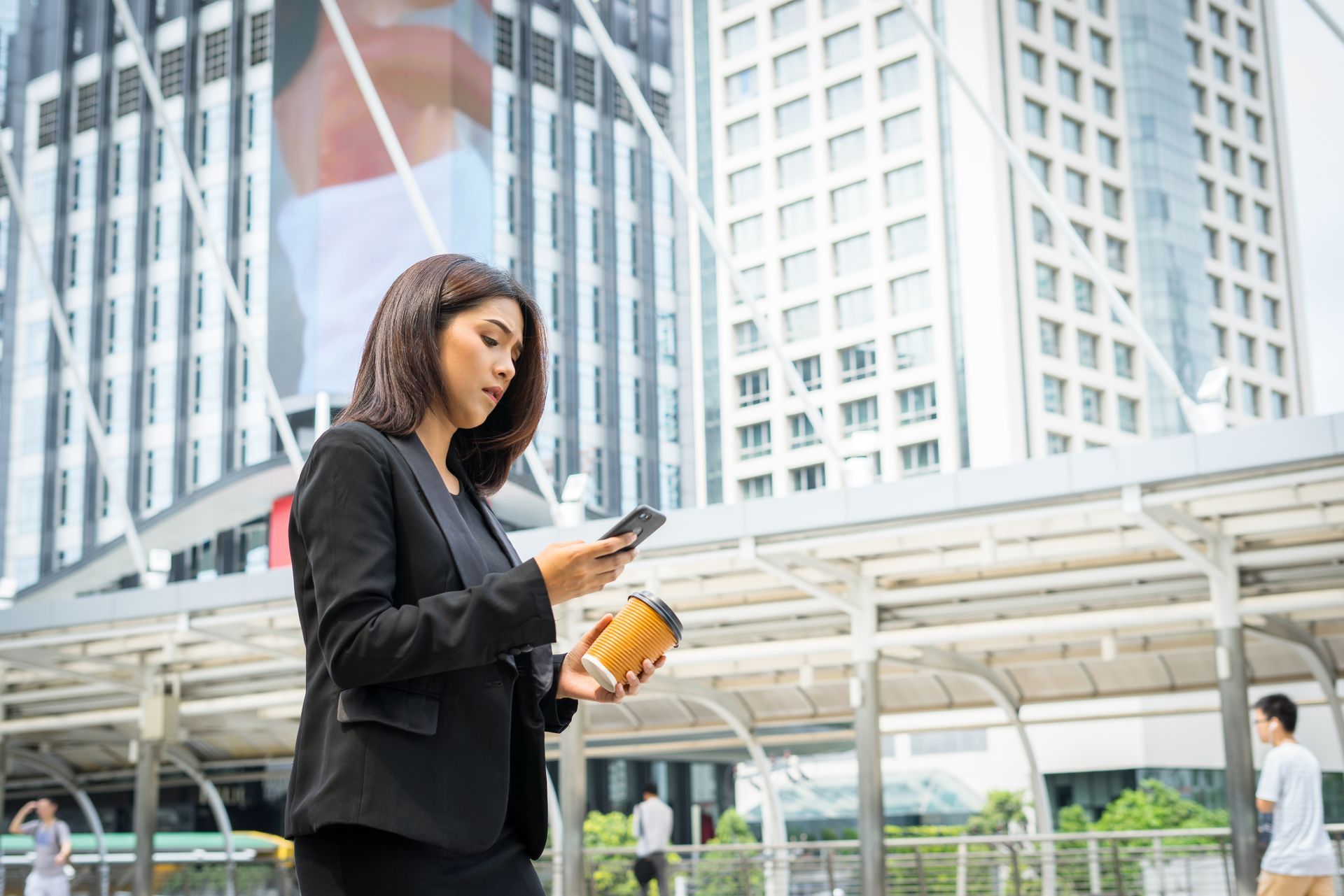 A professional in a black blazer stands outside, checking a smartphone while holding a coffee cup in a city setting.