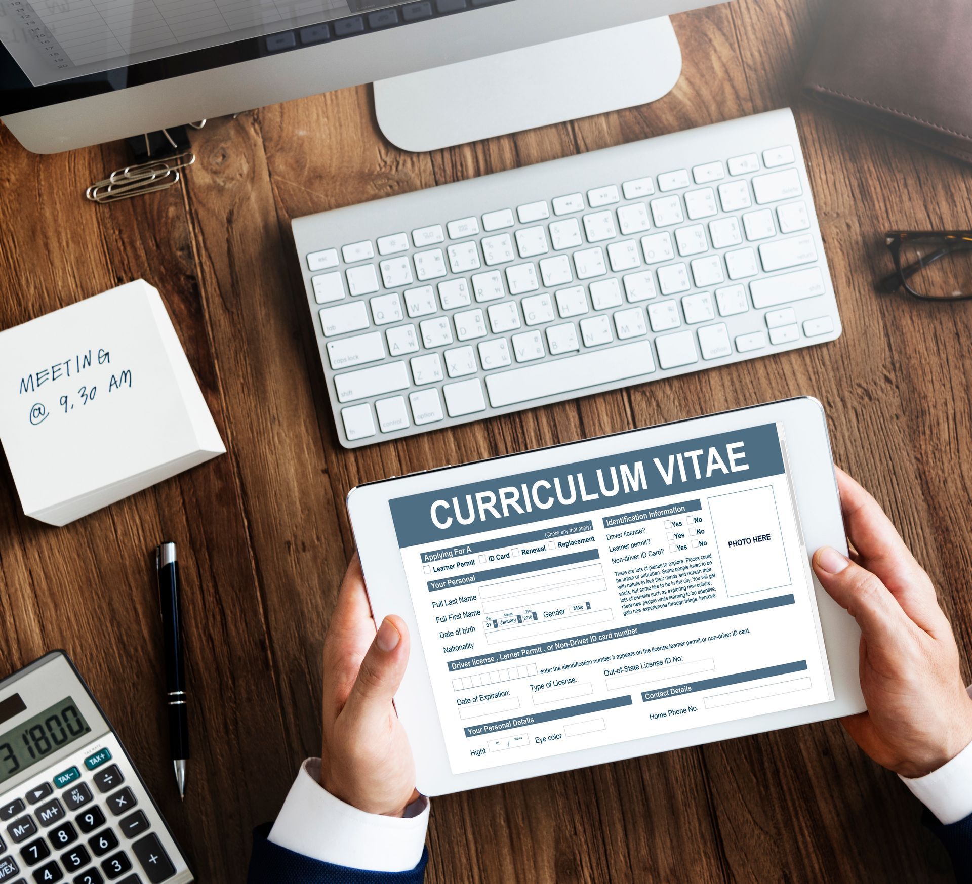 A person holding a tablet displaying a curriculum vitae on a wooden desk with a computer, keyboard, and calculator.