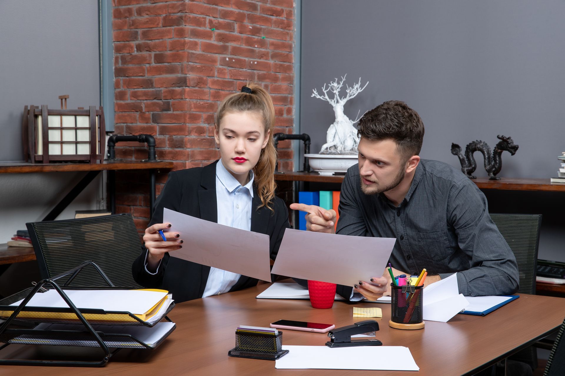 Two people in an office look at documents on a desk, with one gesturing toward the page while they collaborate.