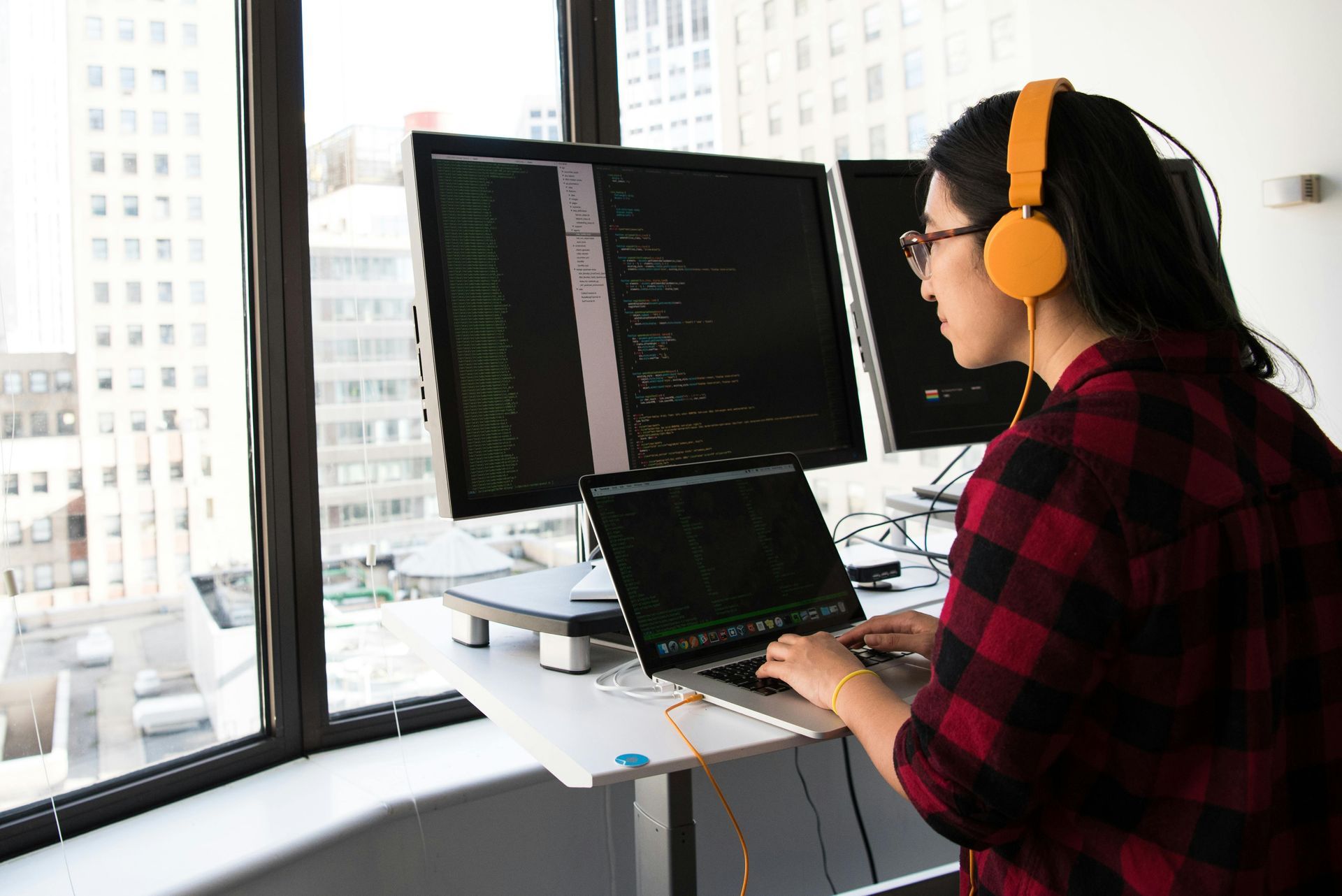 A person wearing yellow headphones works at a standing desk with multiple monitors in a high-rise office.
