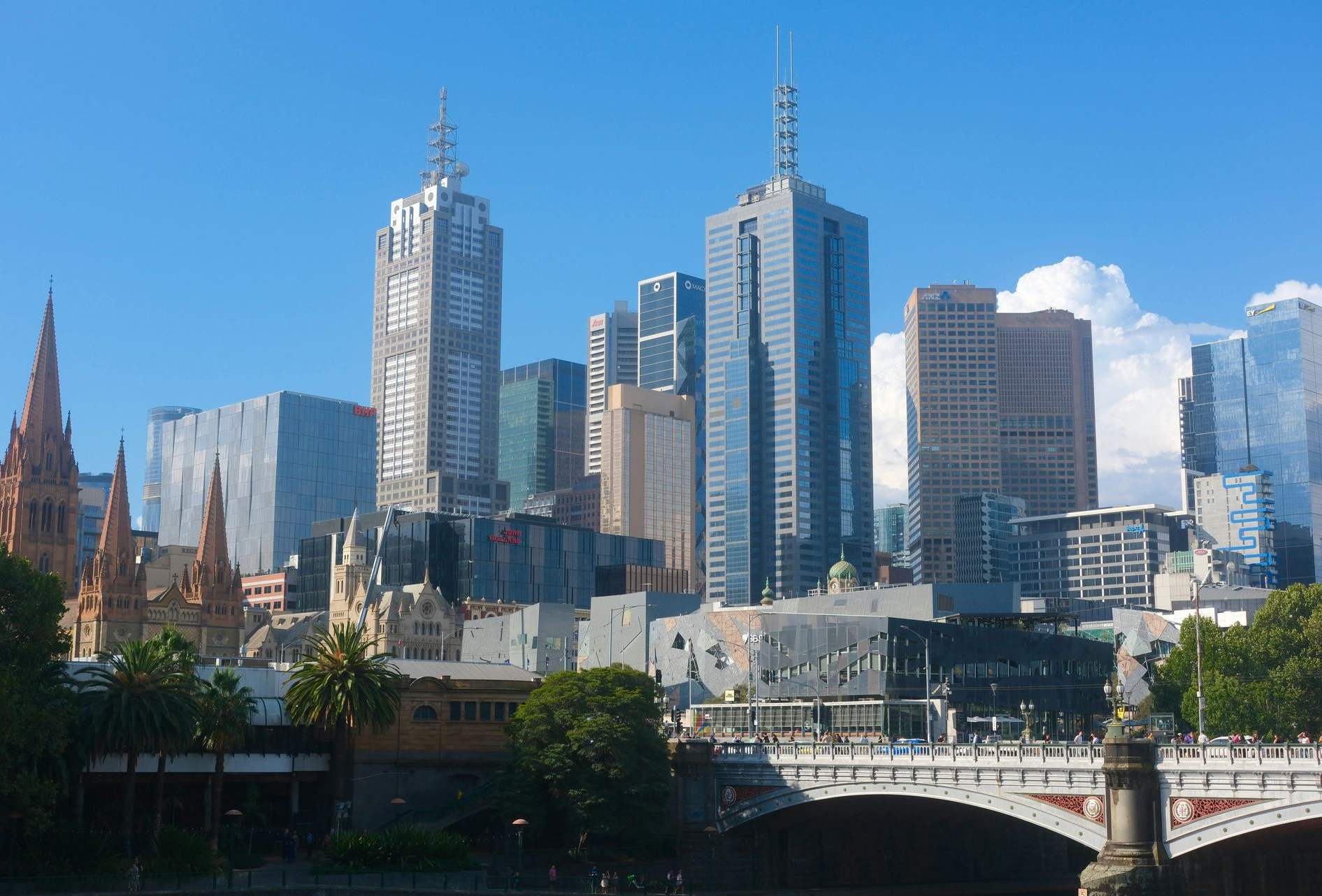 Melbourne skyline with modern skyscrapers against a blue sky, trees in foreground.