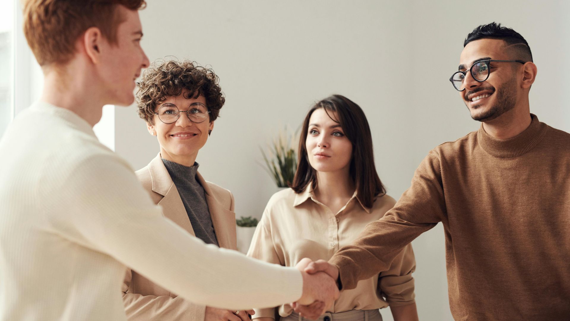 A person with red hair shakes hands with another person, while two others look on in an office setting.