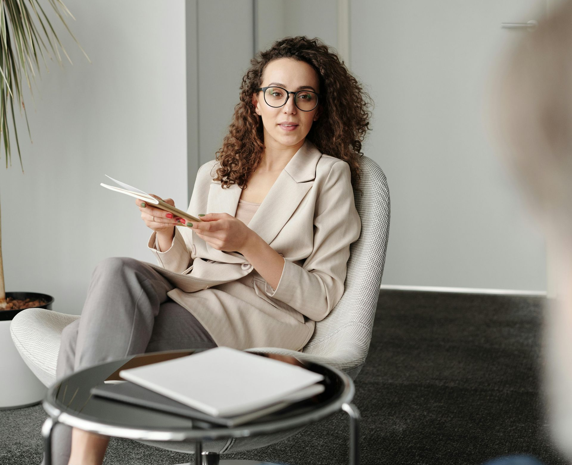 Woman in glasses, beige blazer, and grey pants seated, holding papers, talking, indoors.