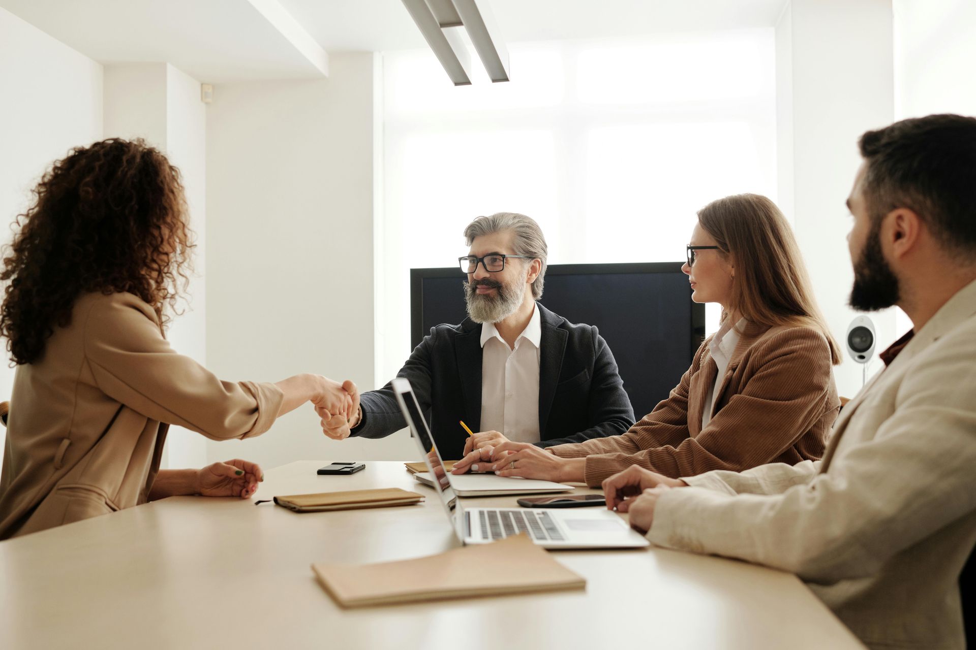 Four people in a meeting, shaking hands. One person is smiling, another looks on, and there are laptops on the table.