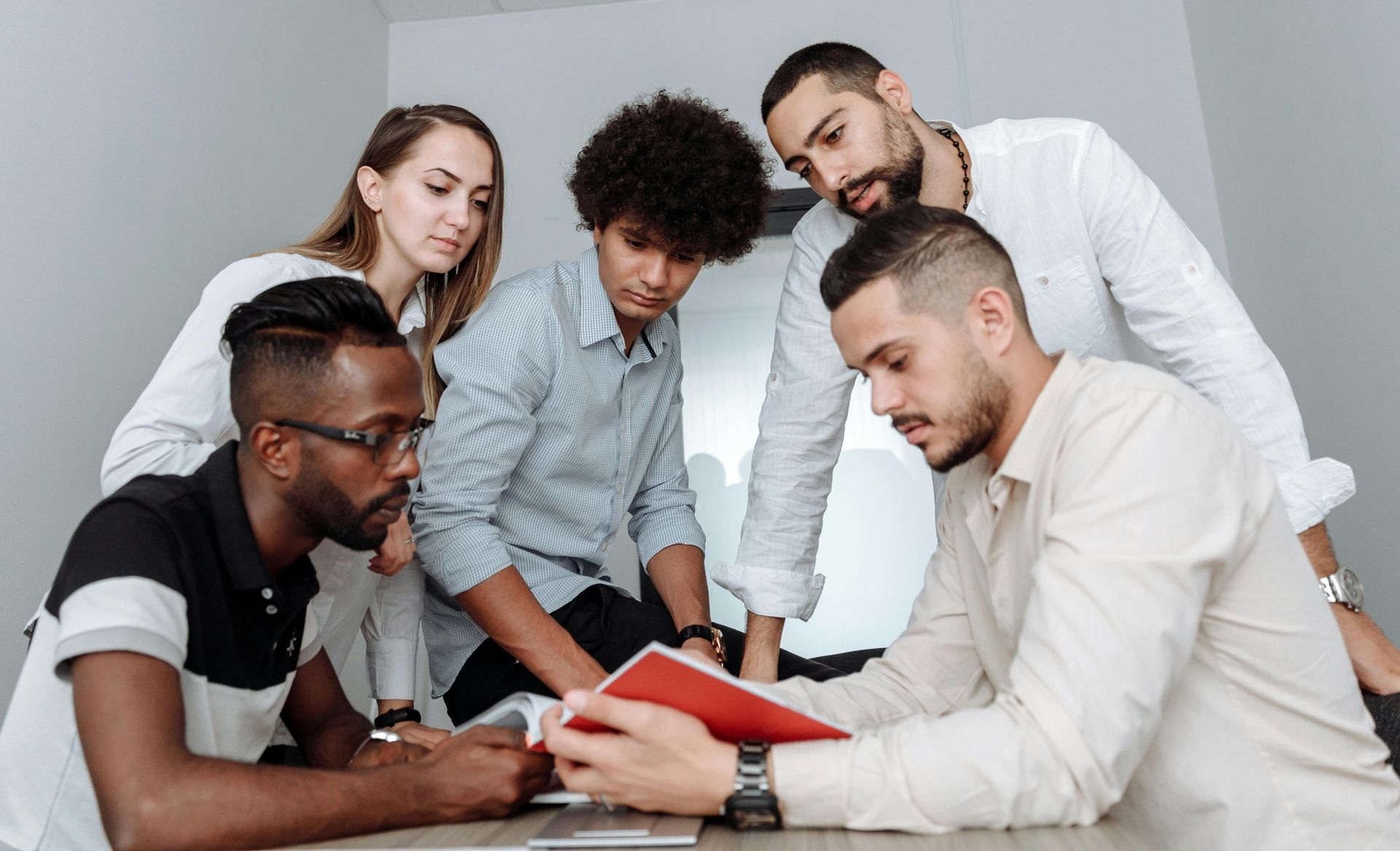 A diverse group of five professionals gathers around a table, focused on a red notebook in a brightly lit office setting.