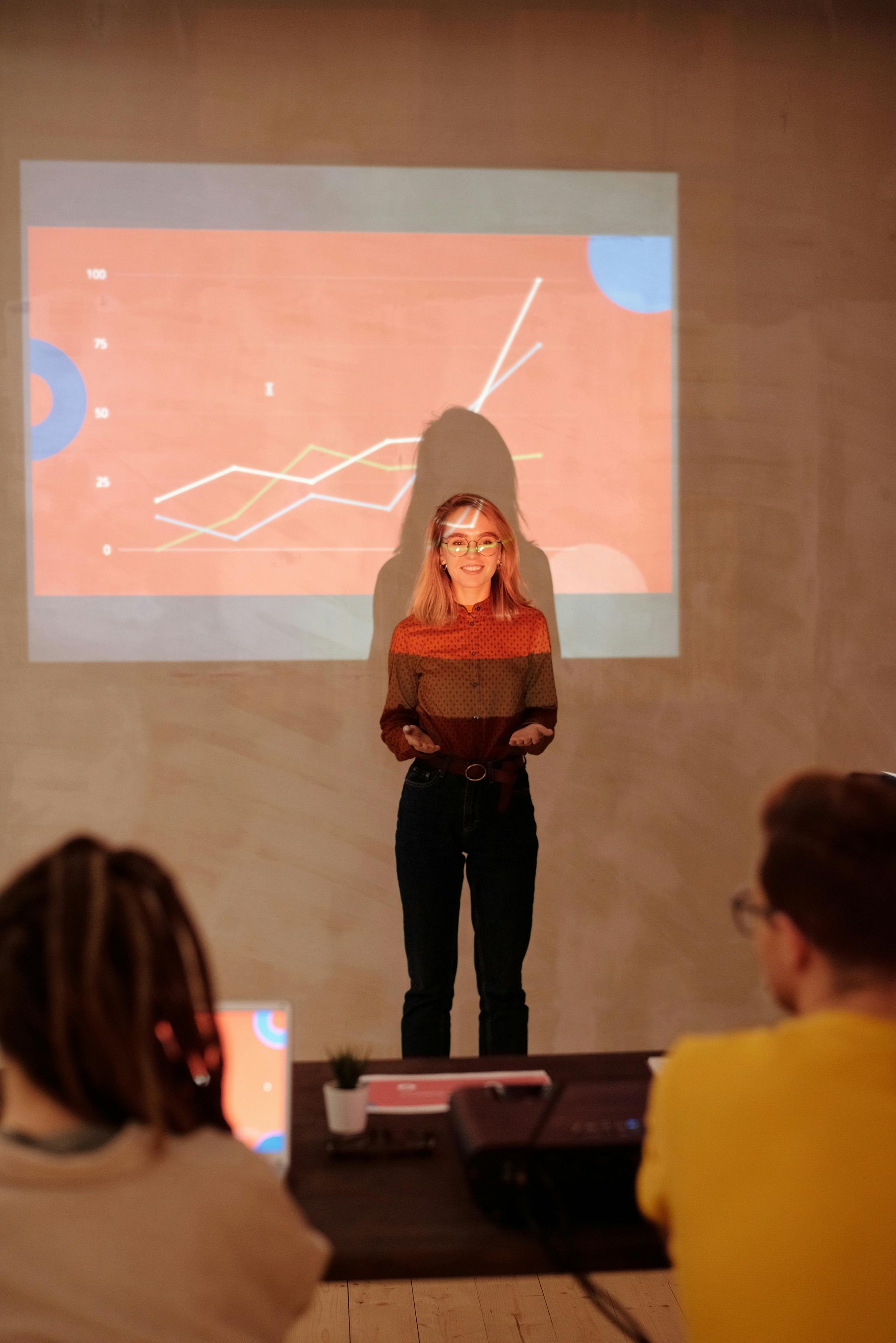 A person stands before a projection screen showing a line graph, giving a presentation to a small audience in a dim room.
