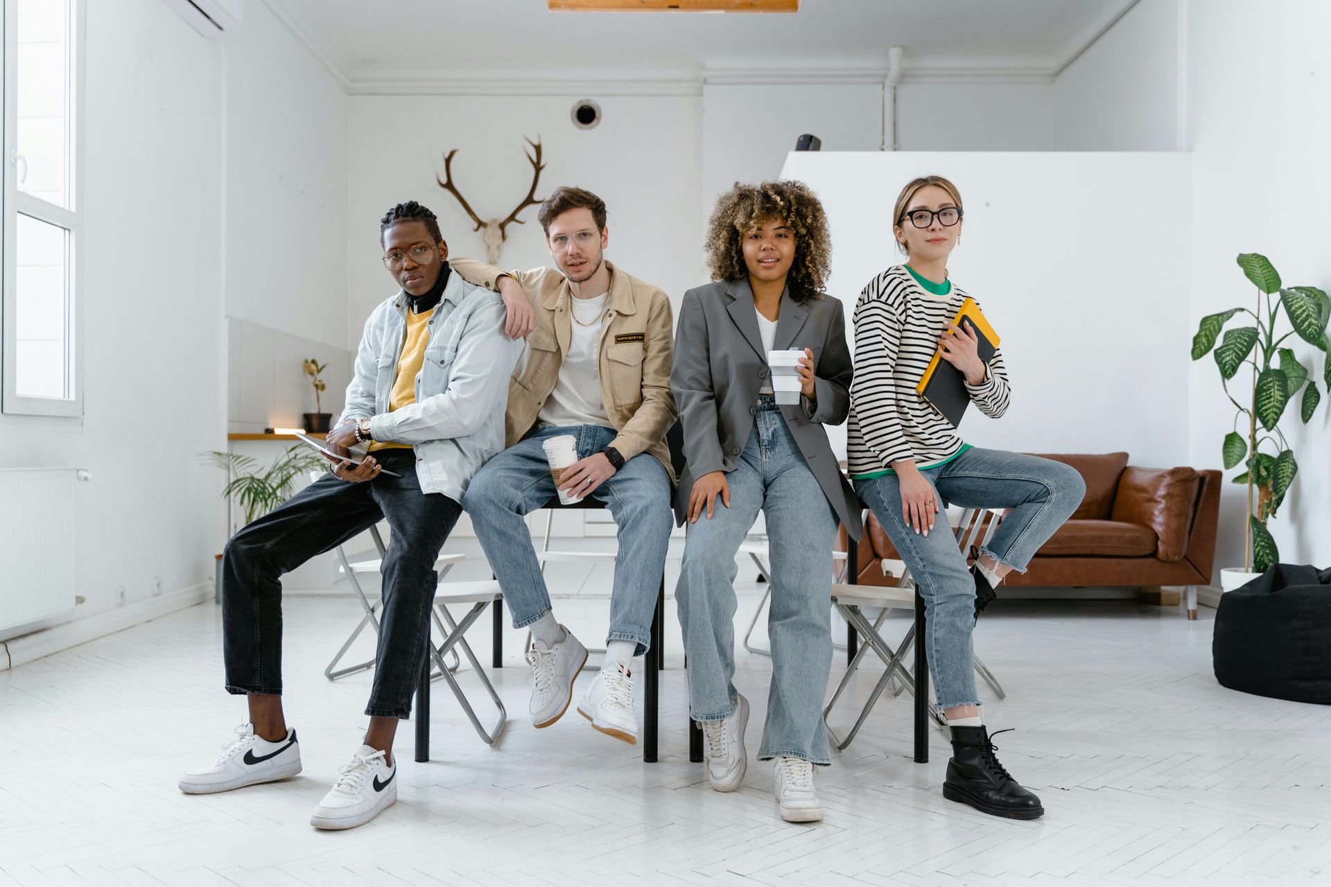 Four people sitting on chairs, looking at the camera. Bright room with a sofa and a plant.