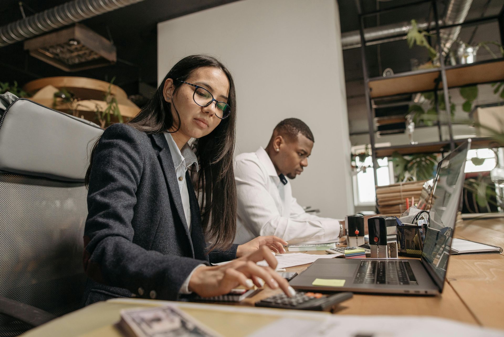 A person in a business blazer calculates numbers on a desk while a colleague works on a laptop in a modern office.