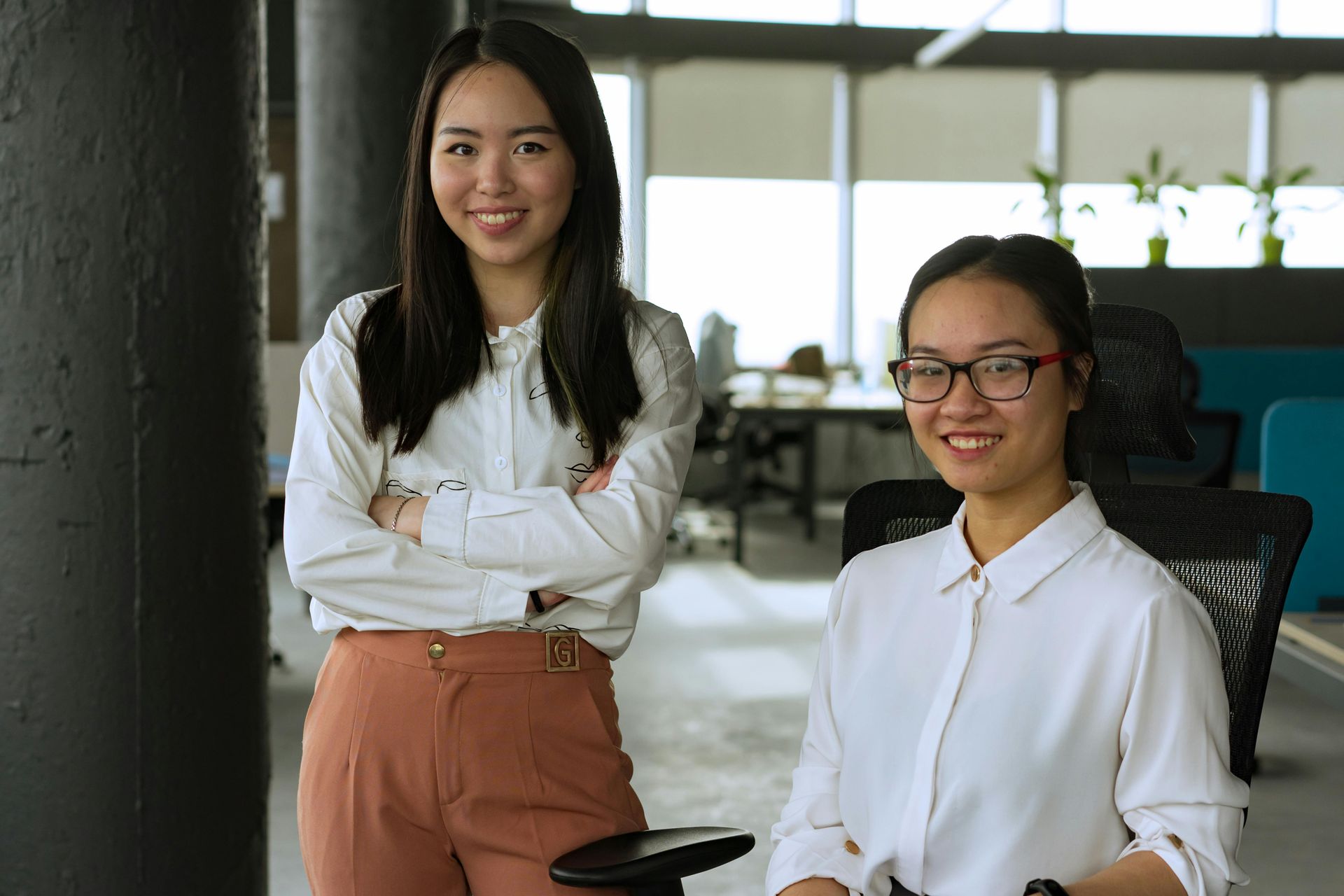 Two smiling people in white shirts pose in a modern, bright office setting, one standing and one seated.