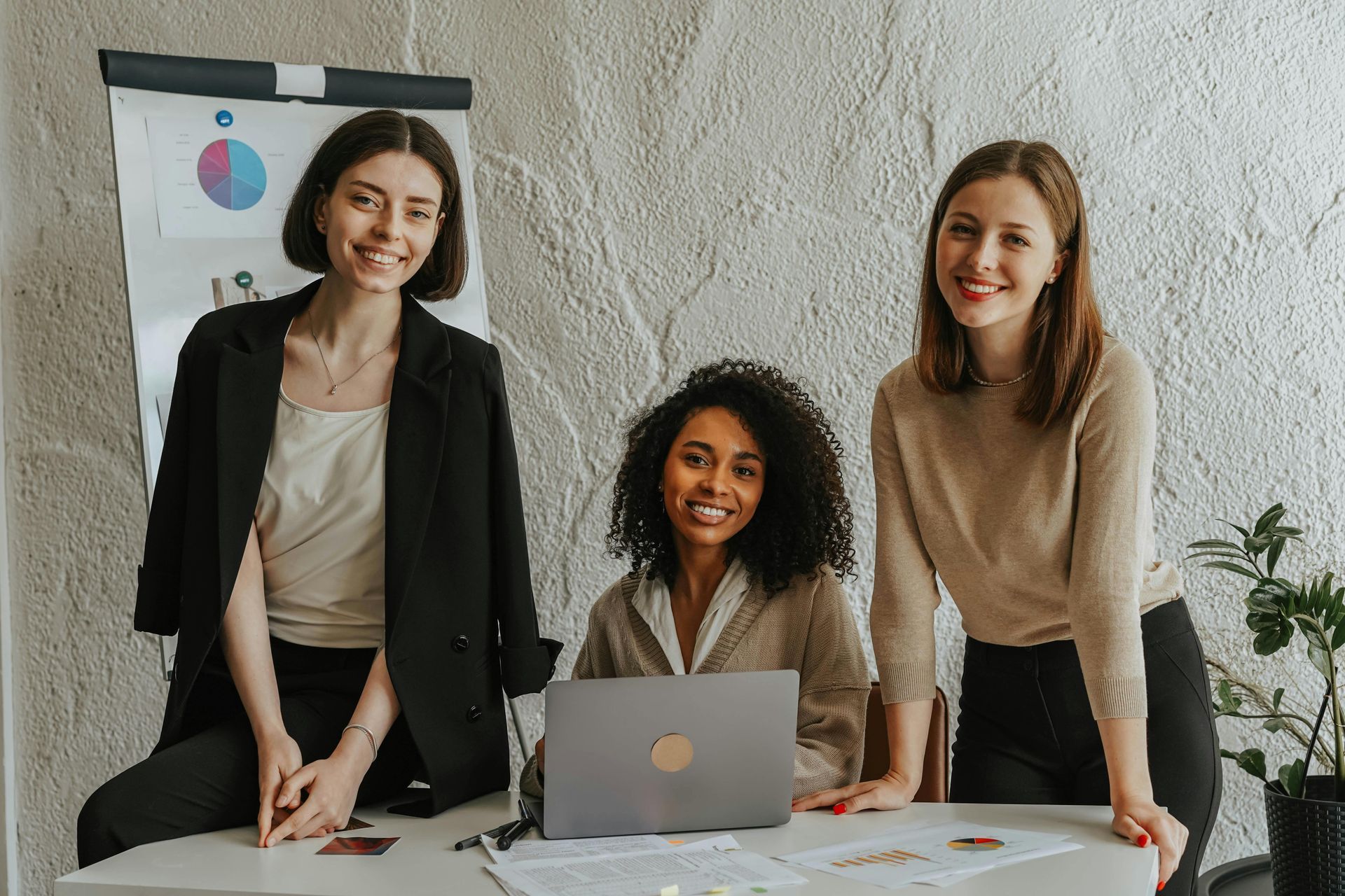 Three women smiling near a desk with a laptop and a whiteboard with a chart in a neutral-toned office.
