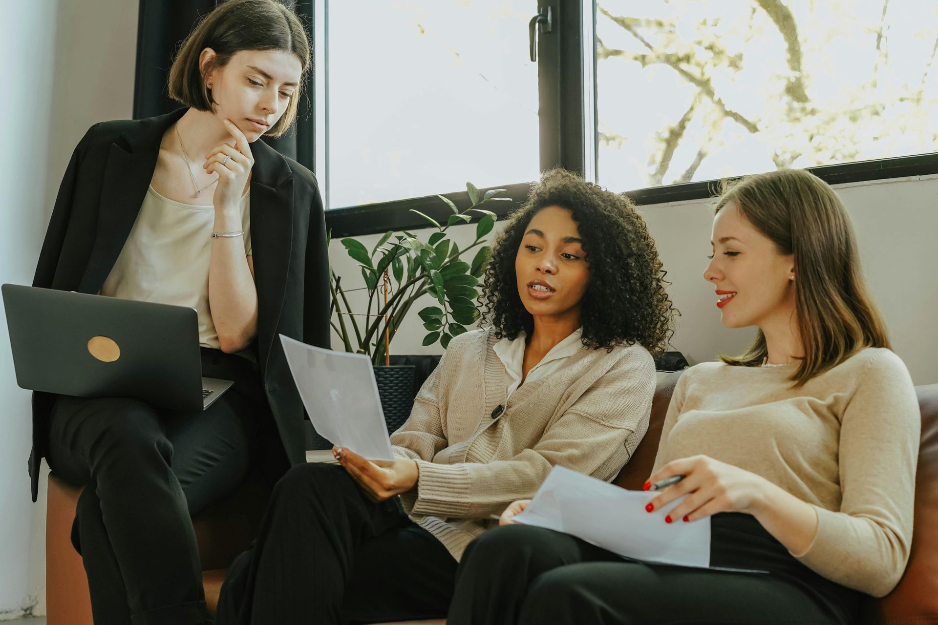 Three colleagues collaborate in an office, reviewing documents and discussing work while seated near a window with plants.