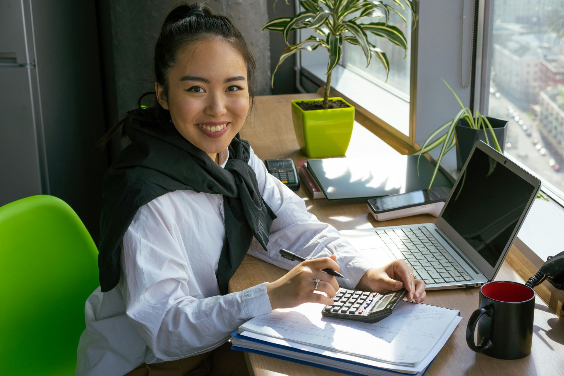 Woman at desk smiles, uses calculator, laptop open, near window with plant.