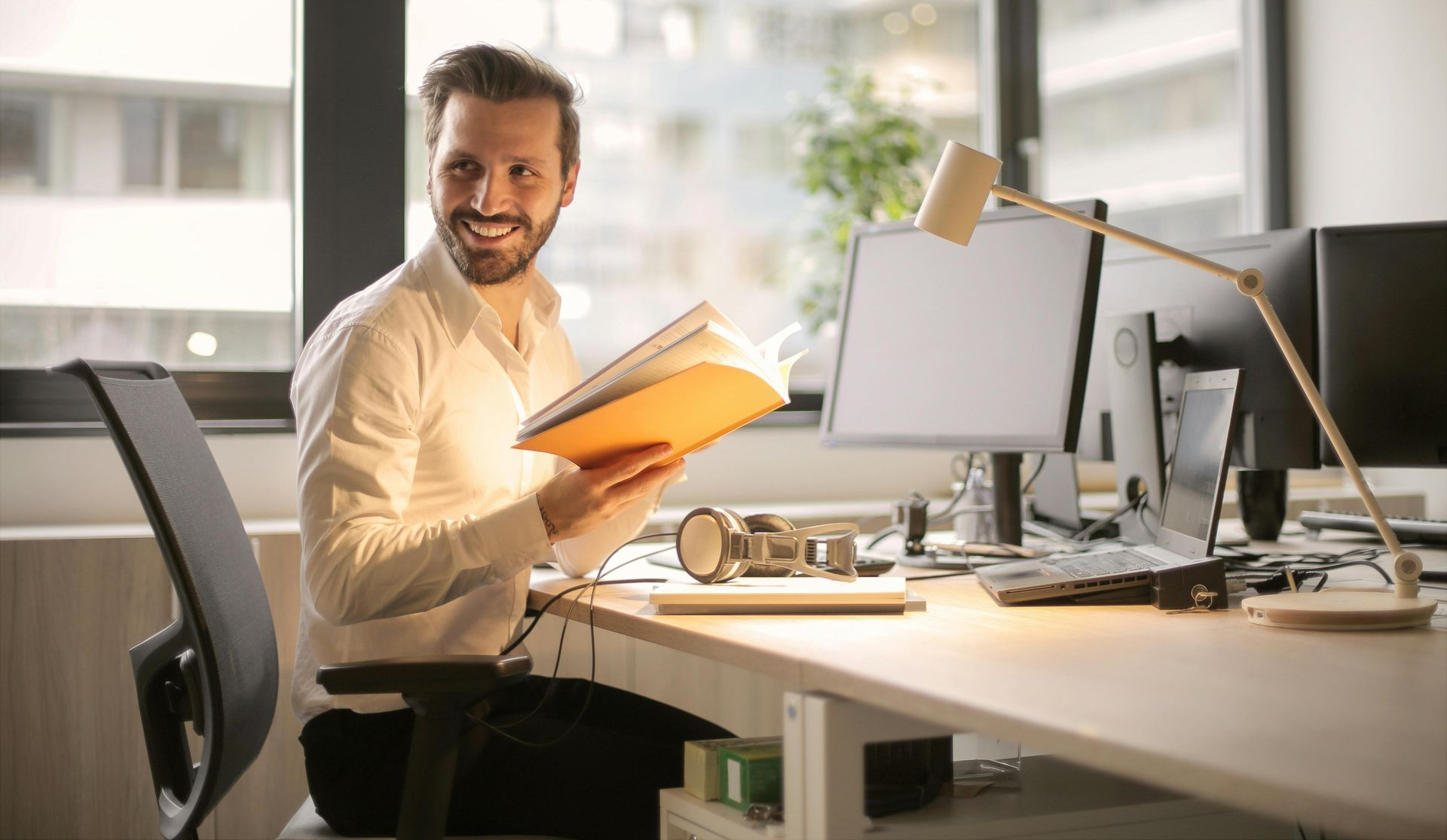 Man smiles, holding orange book at a desk with computer monitors, lamp, and window.