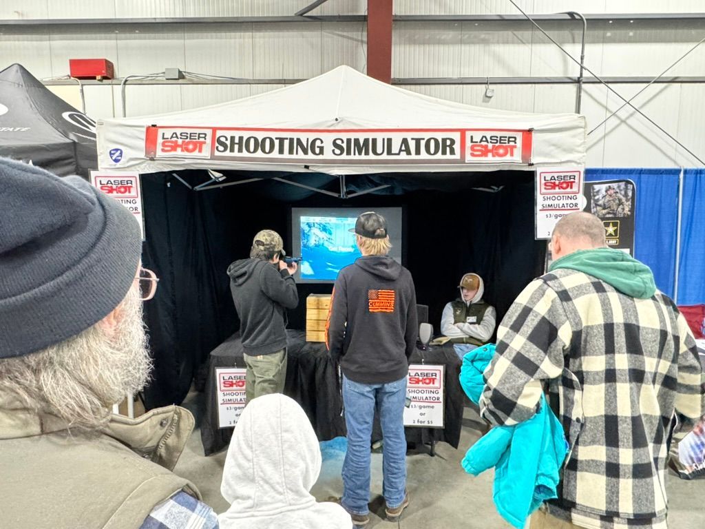 People at a laser shooting simulation booth. Two are aiming at a screen, others watch. Indoors, fair-like setting.