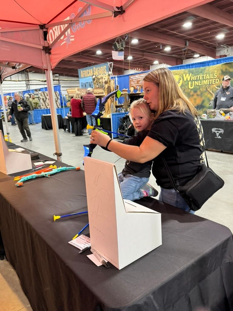 Woman helps a child aim a toy bow and arrow at a target at an indoor event.