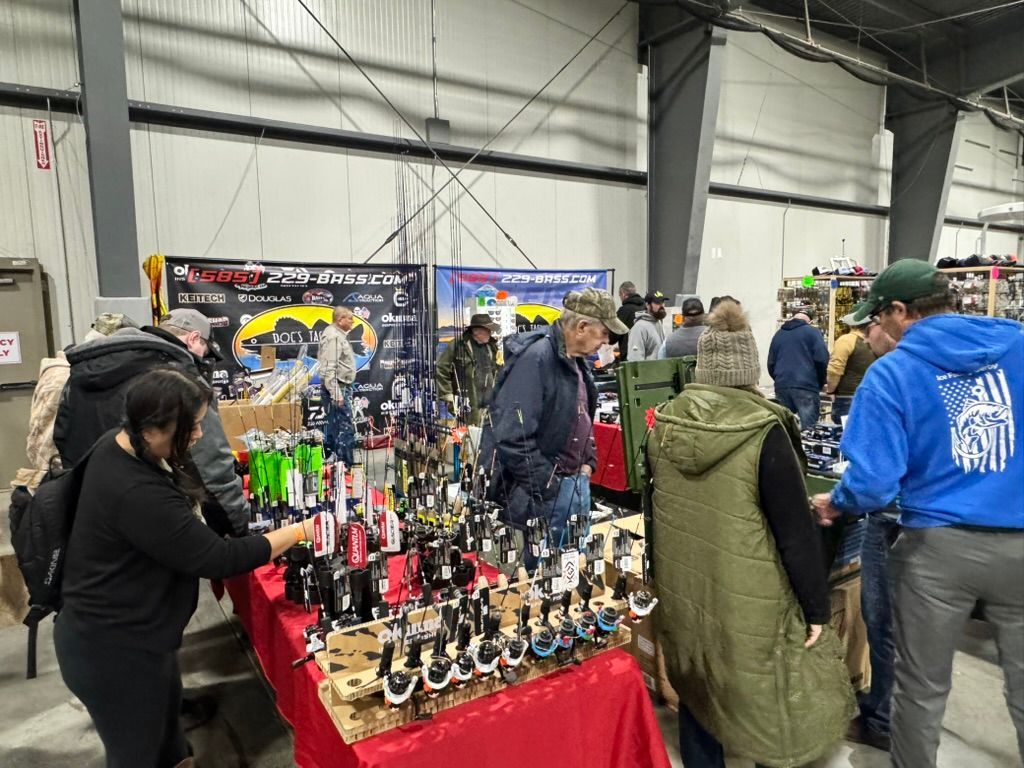 People browsing fishing rods at an indoor vendor fair; red tablecloths, diverse attire, and overhead lighting.