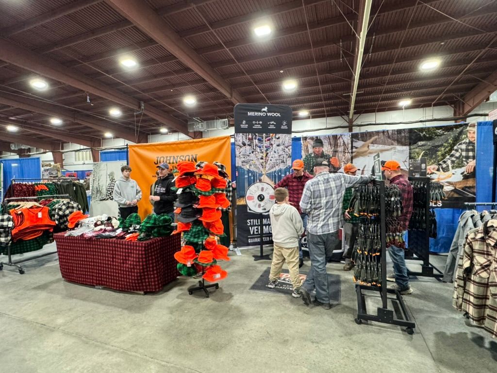 People browsing merchandise at an indoor trade show. Clothing and hunting gear are displayed.