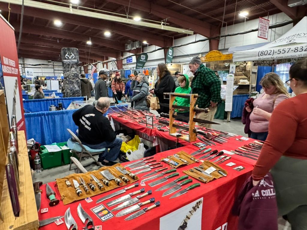 A knife show with vendors displaying various blades. People browse and interact with the displays in a large indoor setting.