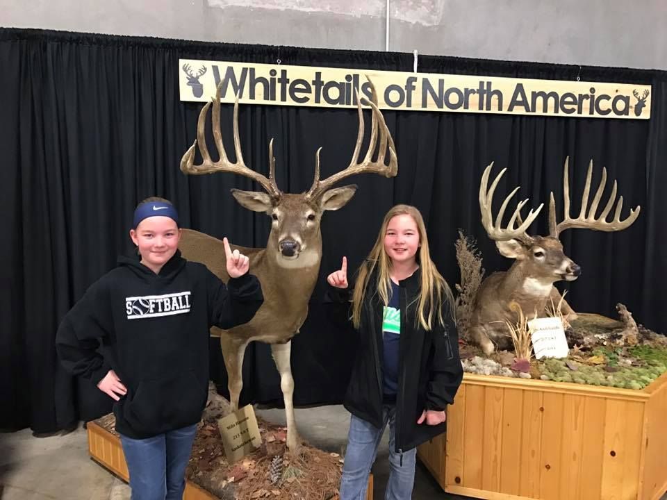 Two girls are standing next to a stuffed deer in front of a sign that says whitetails of north america.
