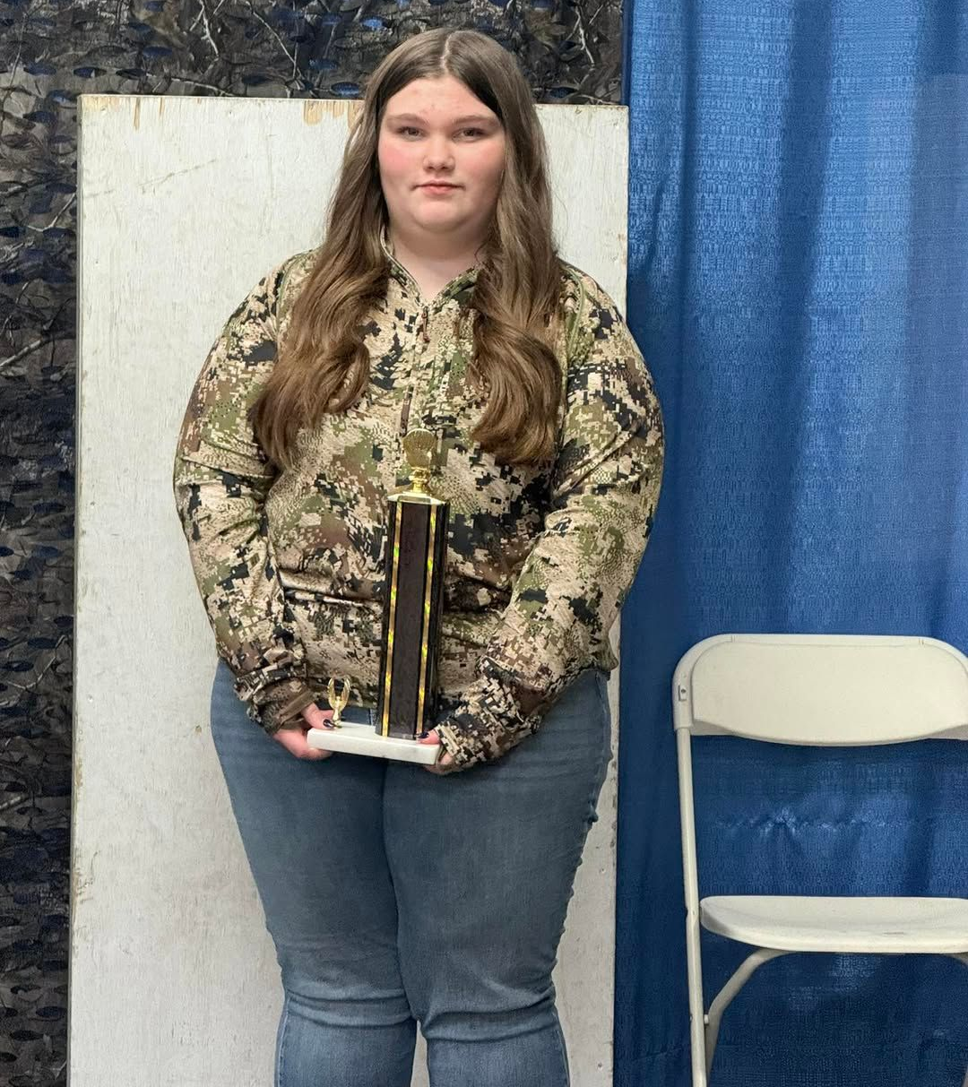 Woman in camo hoodie, holding a trophy, standing in front of a blue curtain.