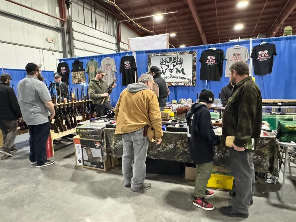 Gun show. People browsing weapons and merchandise at a booth with blue backdrop.