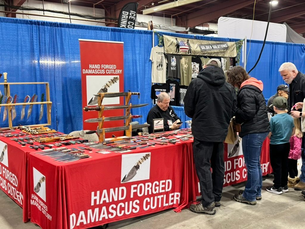 Booth at a show, displaying hand-forged Damascus cutlery. People browse knives on a red table.