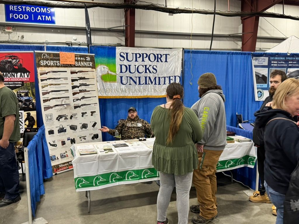 Ducks Unlimited booth at an event. Man in camo, people browsing. Banner says 