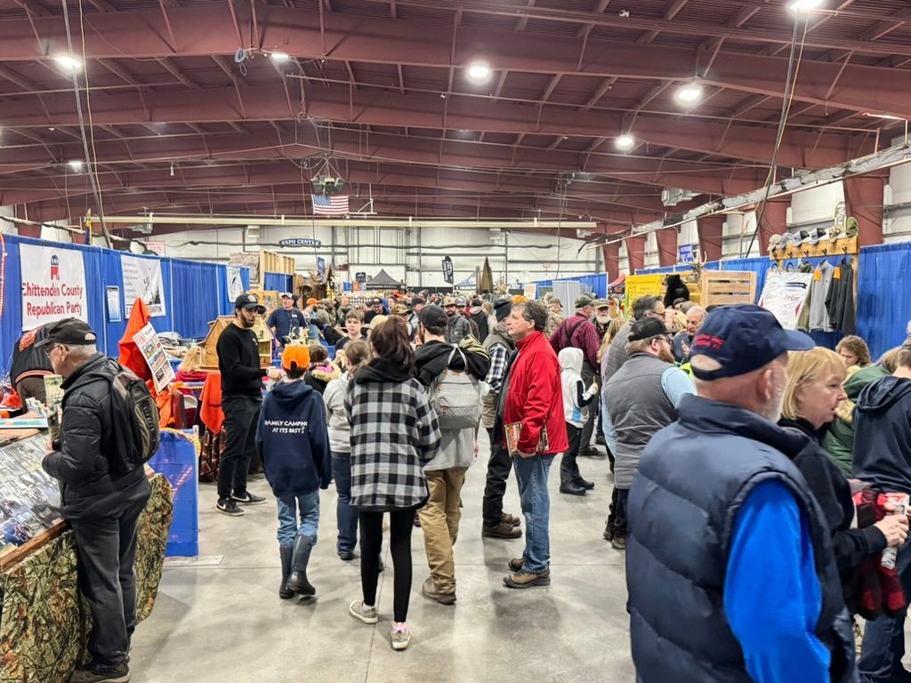 Large indoor event with many people browsing booths, red, blue, and white colors, and items for sale.