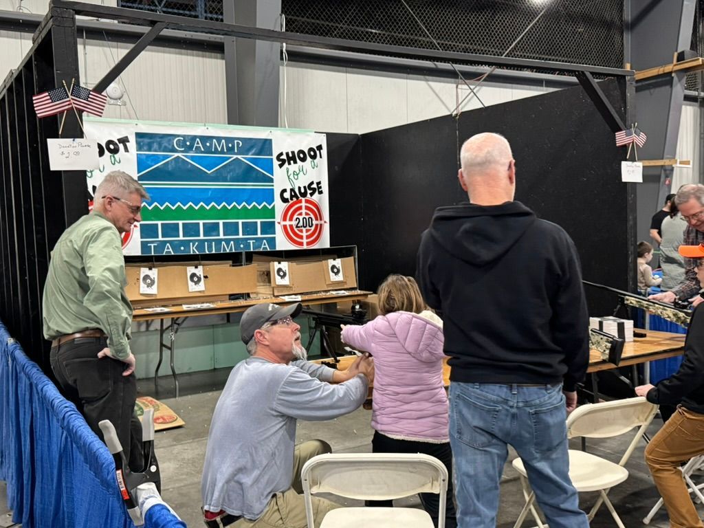 People at a shooting range: one aims, others watch. Target in view. Indoors.
