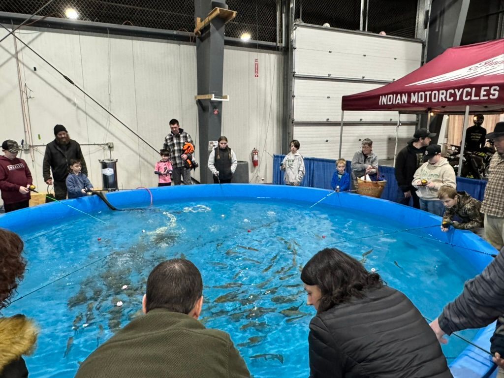 People fishing in a blue kiddie pool at an event, with onlookers and a motorcycle vendor booth in the background.