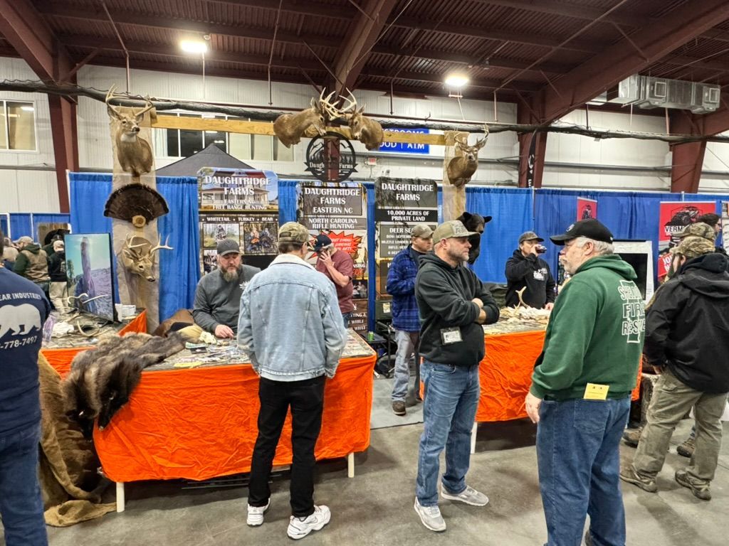 People at an outdoor expo booth, orange tables, deer and elk mounts, indoor setting.