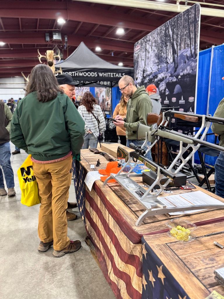 People at a booth, viewing a rifle display. Wooden table with American flag design. Indoor setting, likely an event.