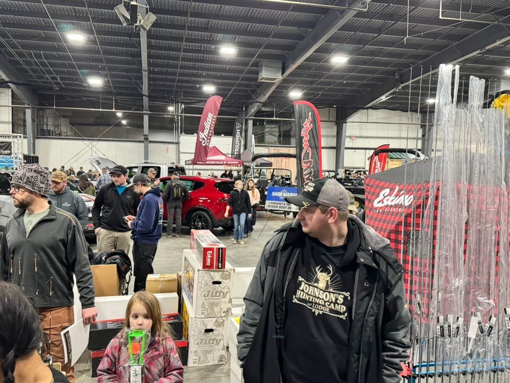 People browsing vendors at an indoor event, possibly a trade show, with fishing gear displayed.