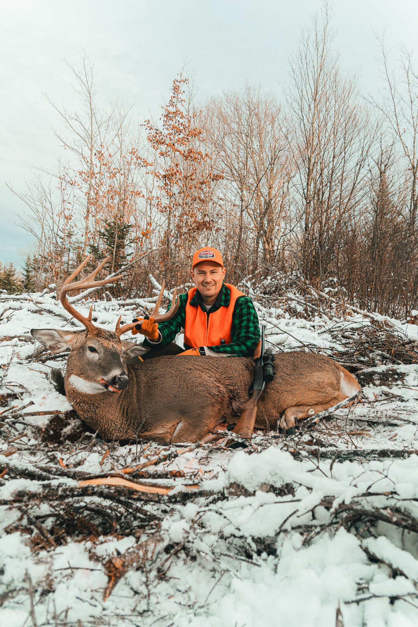 Hunter in orange vest smiles, sitting with a large deer on snowy ground. Forest background.