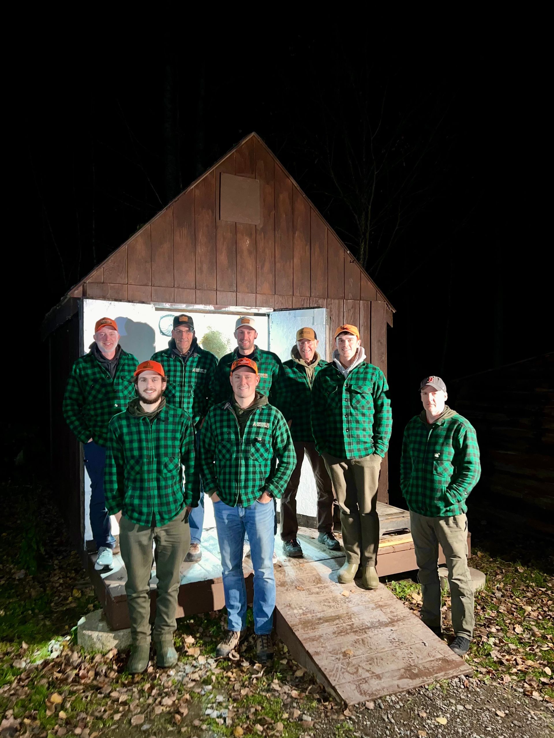 Group of seven people wearing plaid jackets stand in front of a small wooden shed at night.