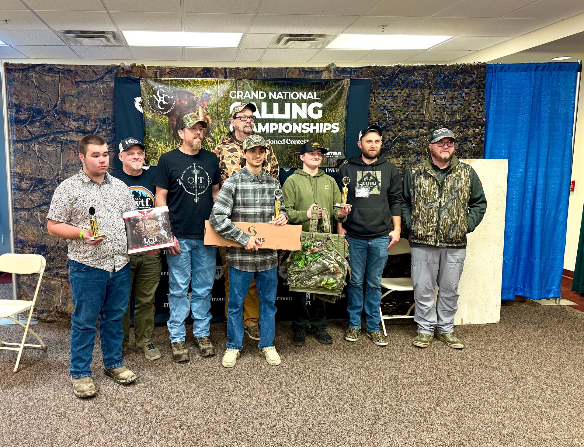 Group of men holding trophies and items at a competition with a camouflage backdrop.