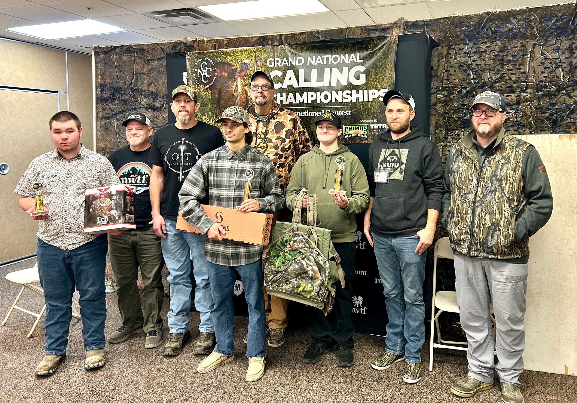 Group of eight men posing with prizes at a calling championship. They stand in front of a banner.