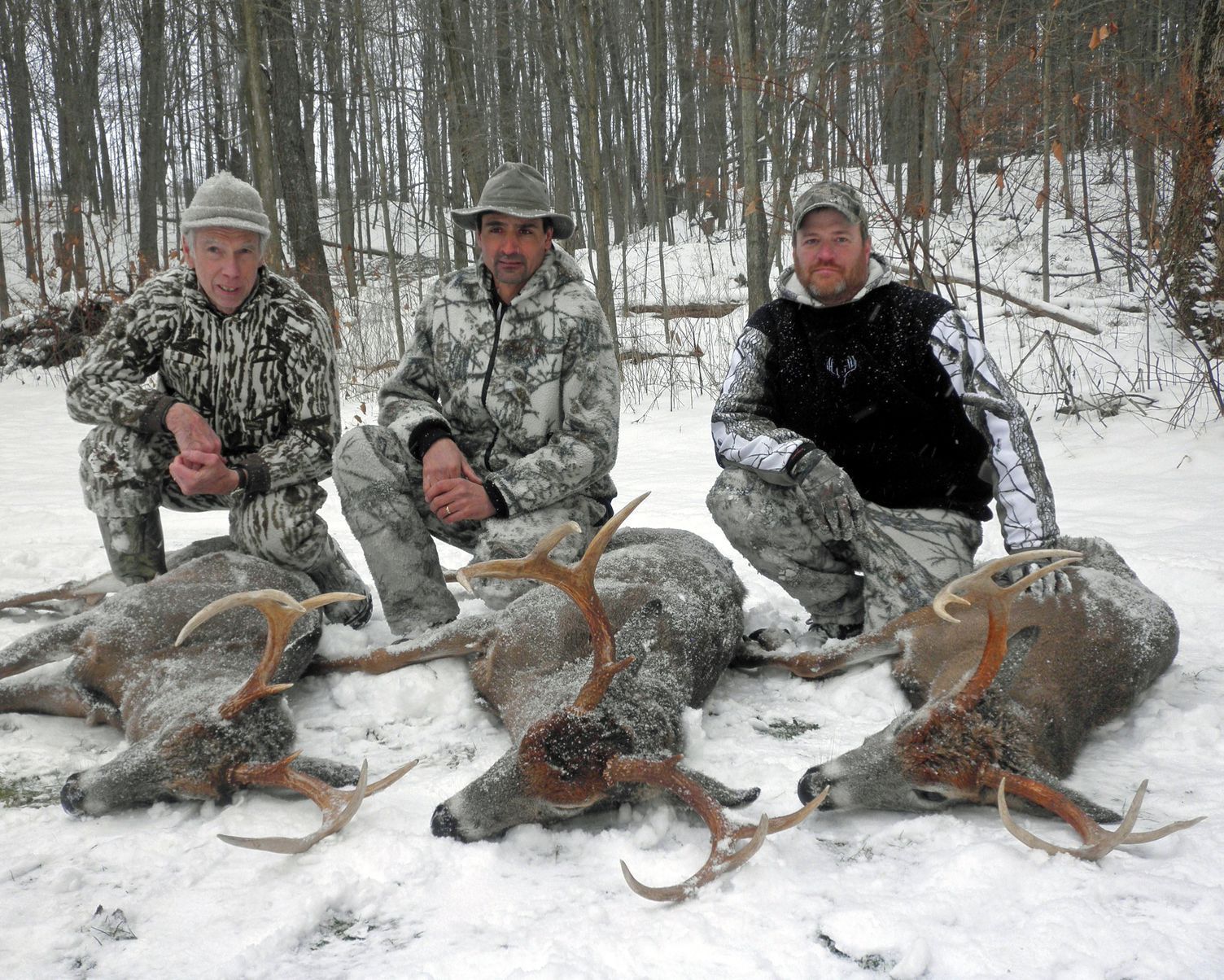 Three hunters kneeling in snow with three deer they have killed.